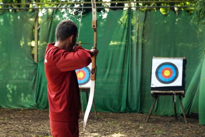 man stands with a bow drawn back, aiming at a target in the distance